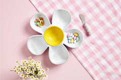 A white Daisy chip platter with a yellow middle for the dip, and knife on the side, on top of a pink table.