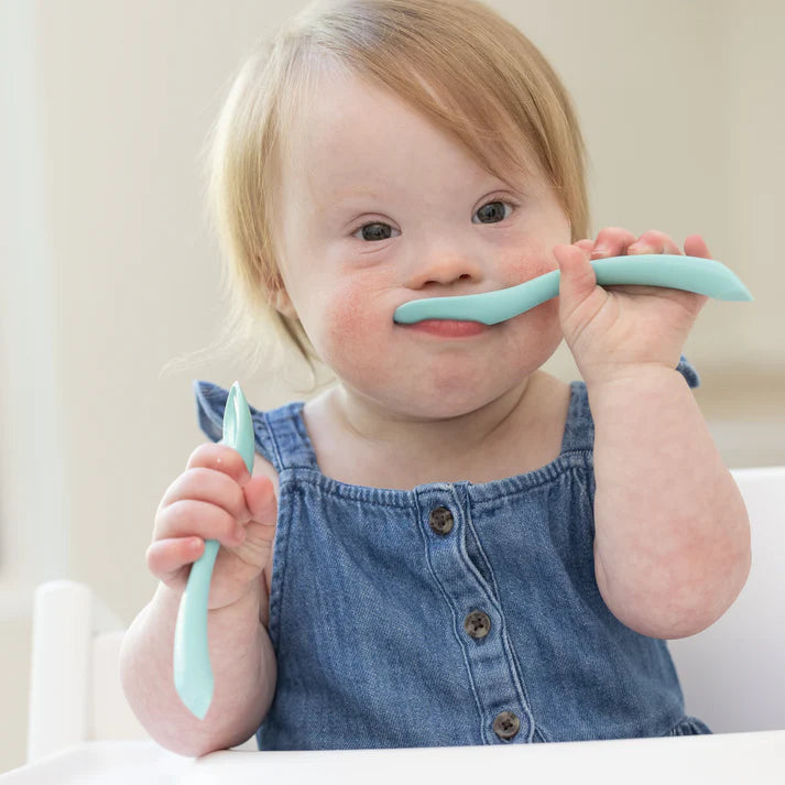 A set of 2 baby spoons in the color teal blue. One spoon reads, "Hello Food I Love You." The other spoon reads, "Baby Got Snacks." This spoon set comes from the brand Bella Tunno.