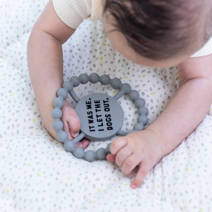 Grey color Happy Teether that reads, "IT WAS ME. I LET THE DOGS OUT" in the middle. The teether is made from silicone, and comes from the brand Bella Tunno.