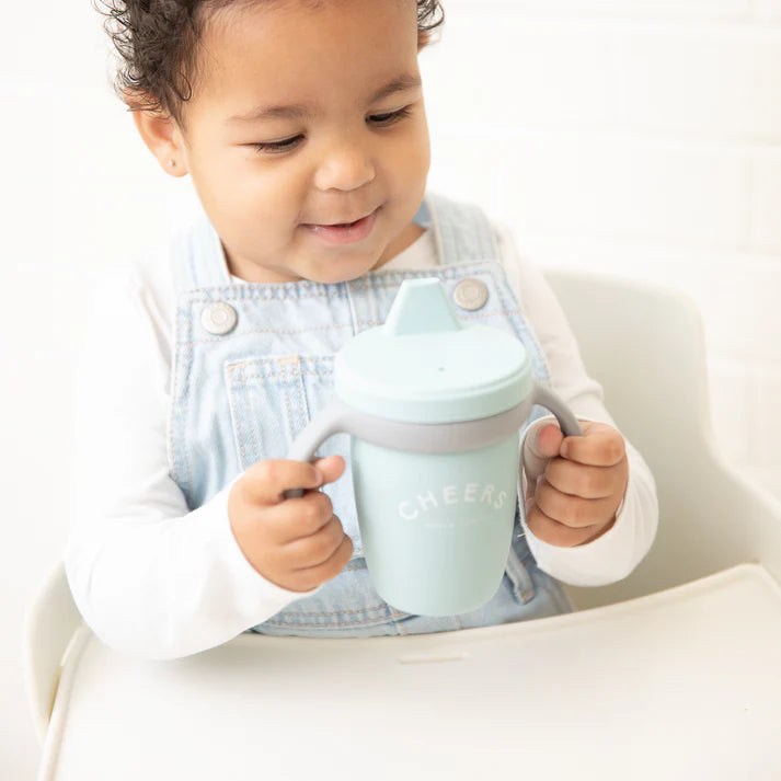 A blue color sippy cup, with gray handles. The sippy cup reads, "Cheers" with the Bella Tunno logo at the bottom.