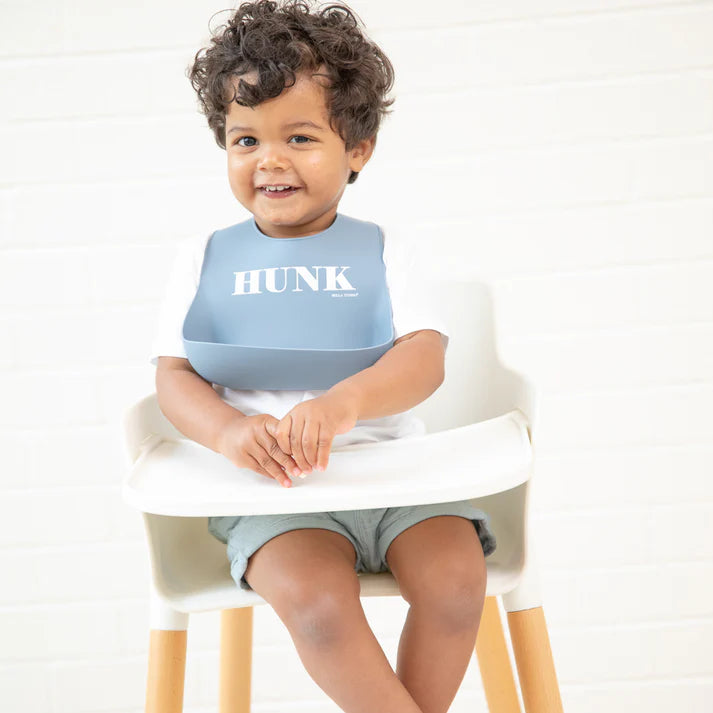 Blue color Wonder Bib that reads, "HUNK" with the Bella Tunno logo below it. The bib is adjustable in size with 4 snaps that easily fit around the neck. The Wonder Bib is designed out of silicone, and is made by Bella Tunno.