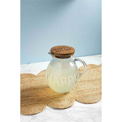 A glass drink pitcher that reads "Happy" on the body, with a brown lid on top that reads, "HAPPY HOME." Sitting on a brown place mat, with a blue wall behind it.