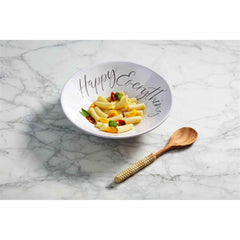 A white bowl that reads, "Happy Everything" sitting on a counter top full of pasta.  With a wooden spoon with a rattan-wrapped handle sitting beside the bowl.