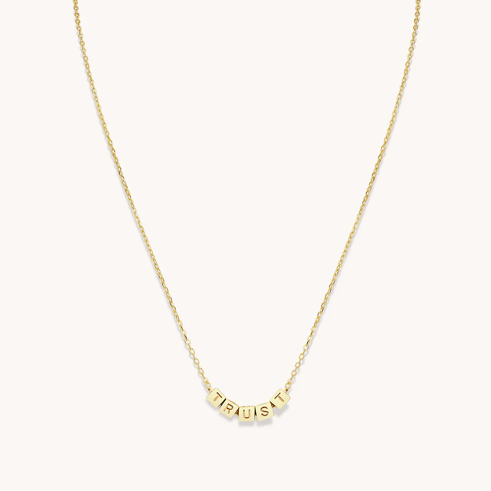 A gold chain necklace from Little Words Project. The necklace includes gold beads that read, TRUST.