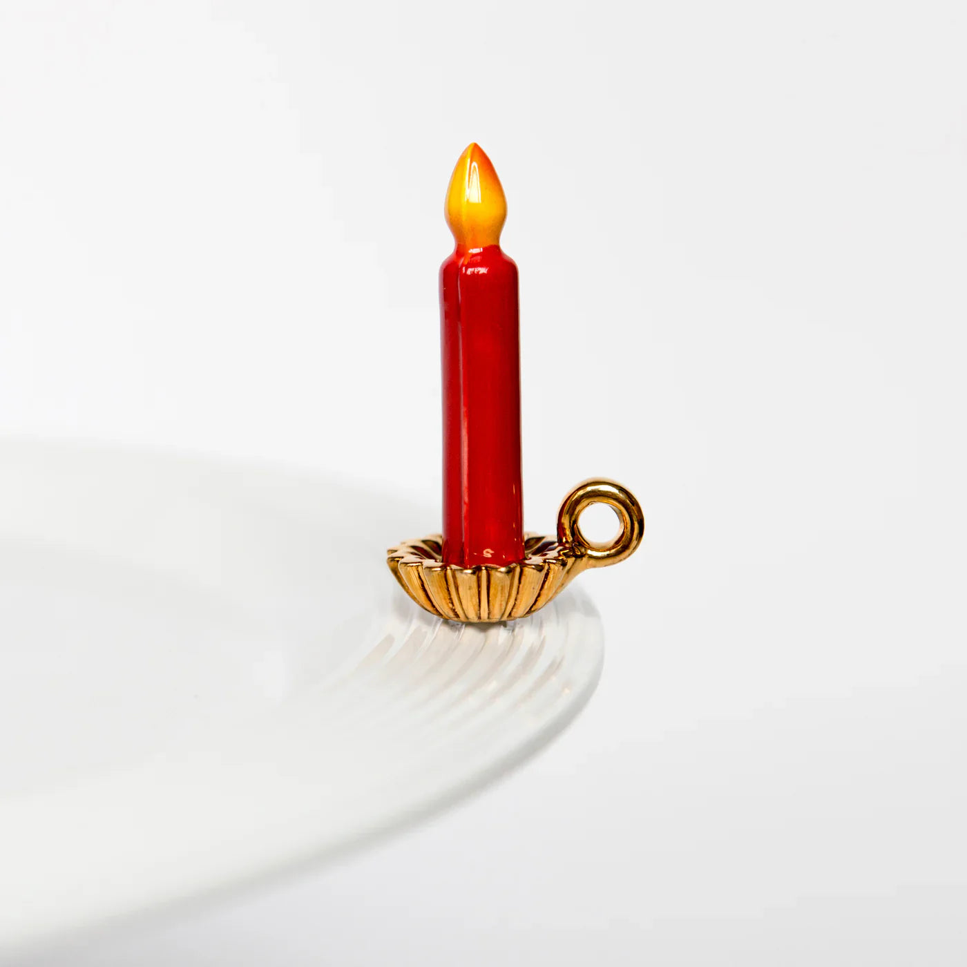 Nora Fleming Light The Way Mini decor item. A red candle with an orange burning flame that sits in a gold holder. The mini offers a stem at the bottom so you can place it onto your favorite platter.