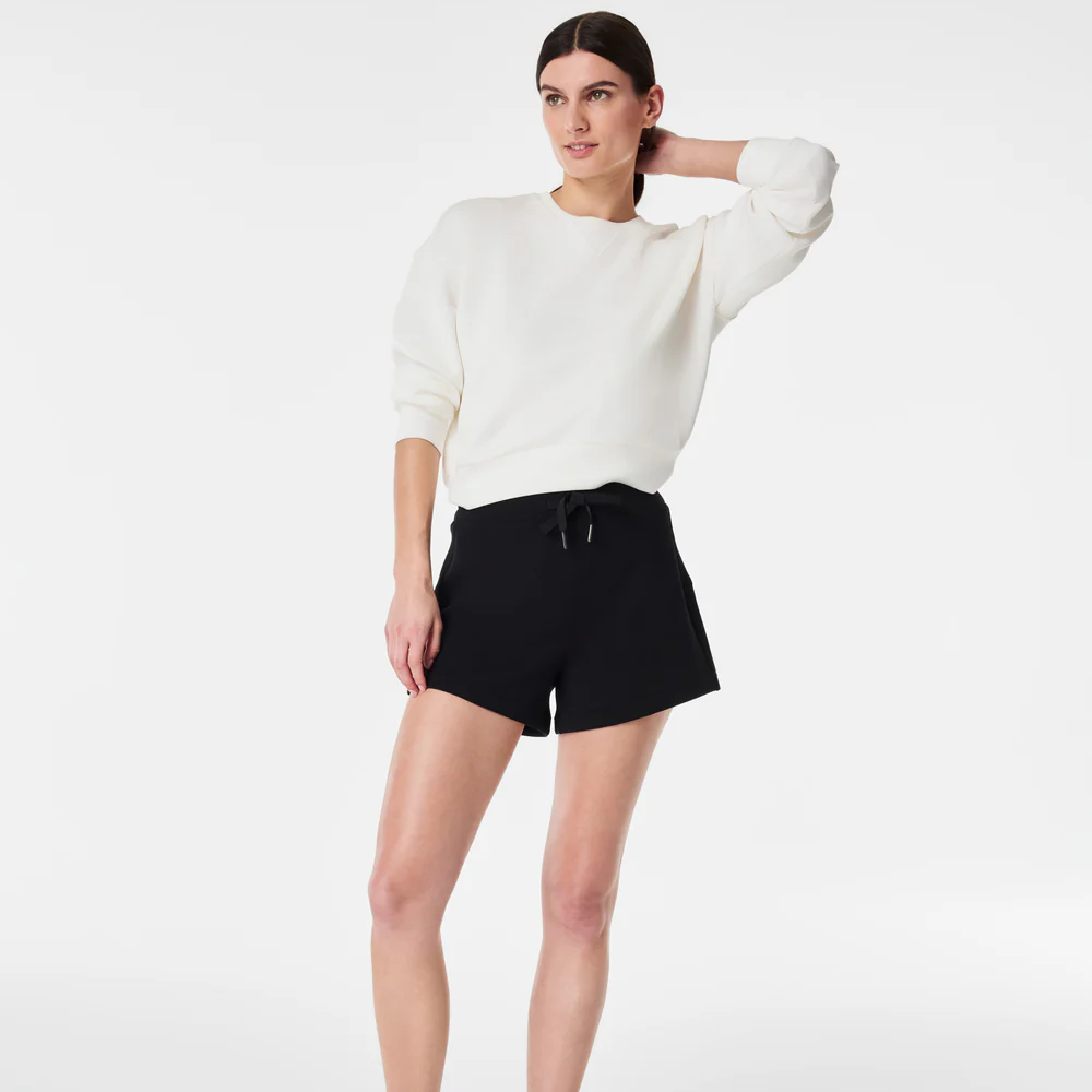 A person stands with one hand resting on their head, wearing a white sweater and black shorts against a plain white background. The person is wearing a black pair of SPANX Airessentials Shorts.