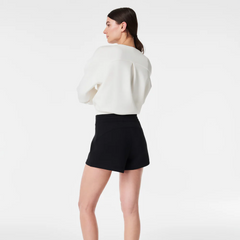 A woman with long dark hair is standing with her back turned, wearing a white Airessentials sweater and black airessentials shorts against a plain white background.