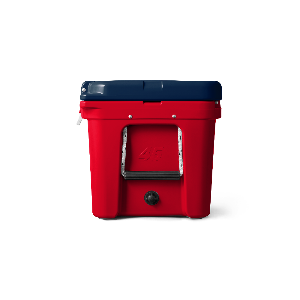 A side view of the limited edition YETI Tundra 45 in Patriotic Red, White, and Blue. The top lid is navy blue, the base of the cooler is red, and the handles on the side are white, with a black hand grip. There is a number 45 engraved into the side of the cooler, also in the color red. The water drain on the cooler is the color black.