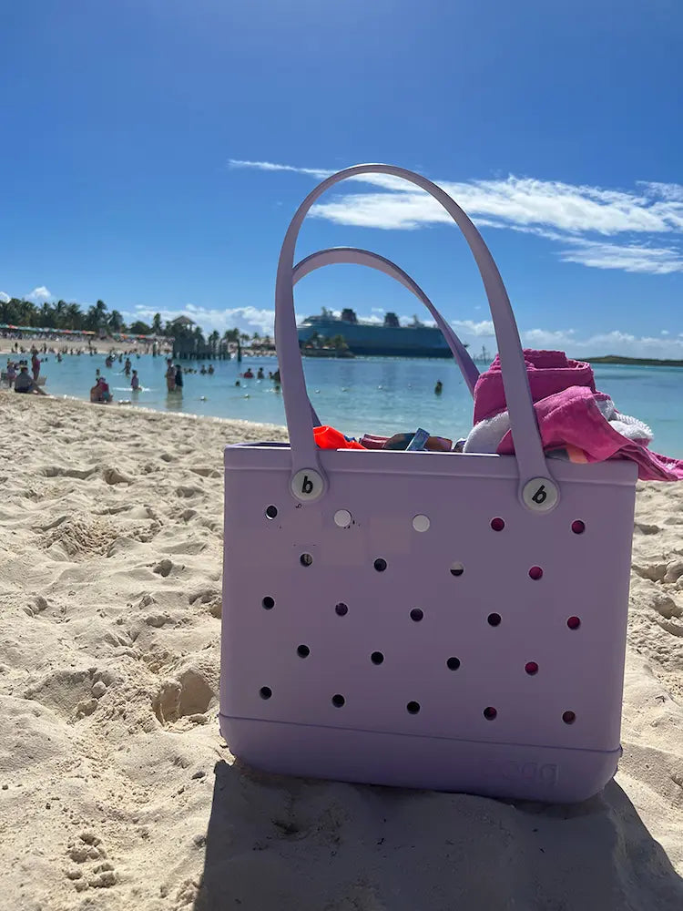 A baby Bogg® Bag chillin' in the sand, full of beach supplies with a curse ship in the background. (This could be you, buy a Bogg® today!)