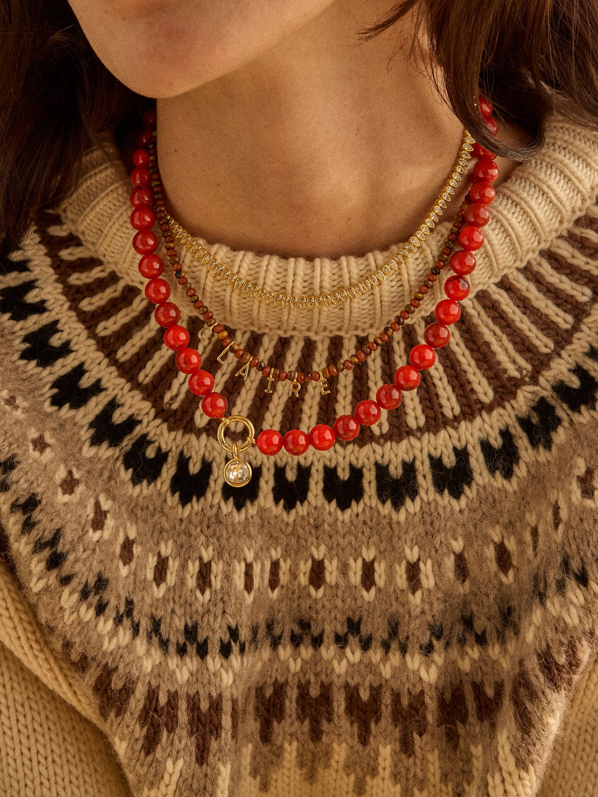 Model wearing the red BaubleBar Janette Beaded Ring Necklace layered with other necklaces over a patterned knit sweater.