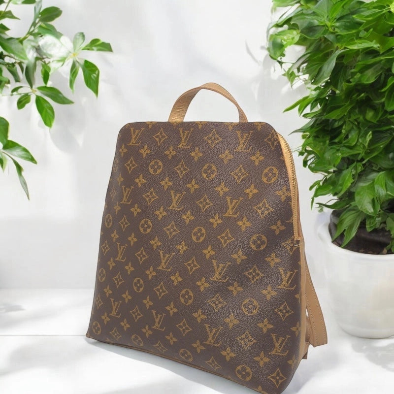 Beaudin Riley bronze backpack with full monogram front displayed beside plants