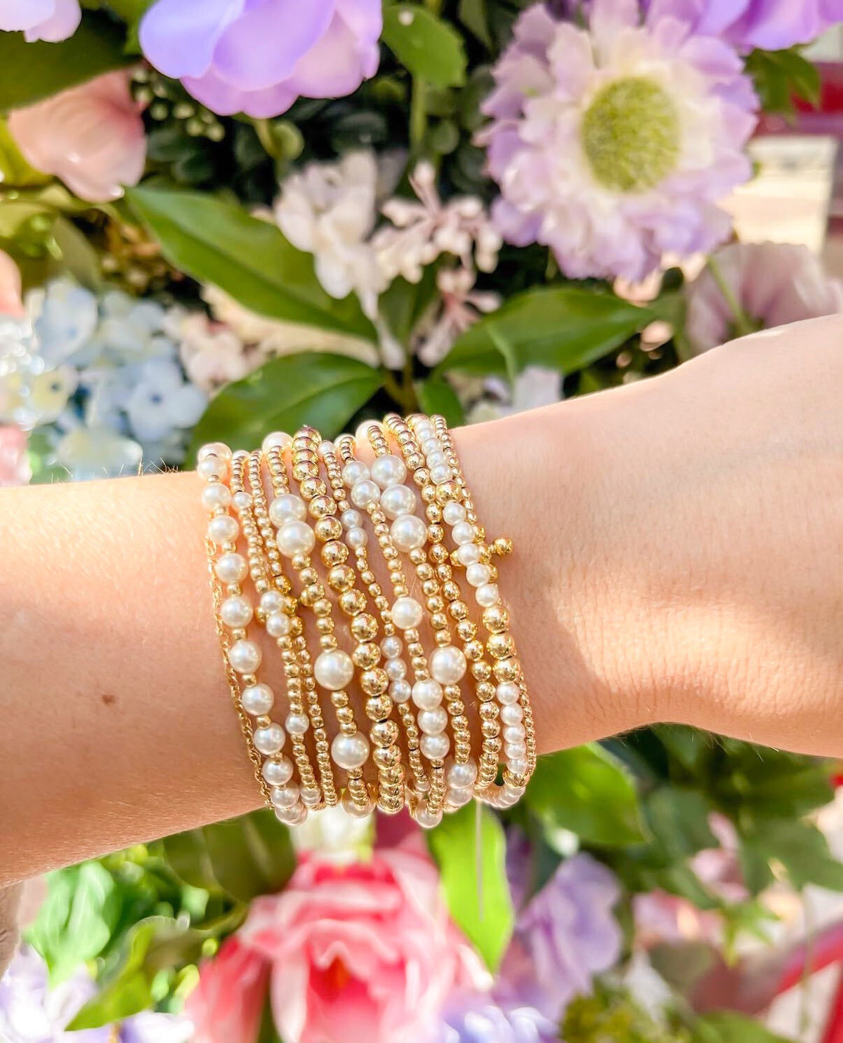 A close-up of a person's wrist wearing a stack of gold and pearl beaded stretch bracelets, set against a soft-focus background of colorful flowers.