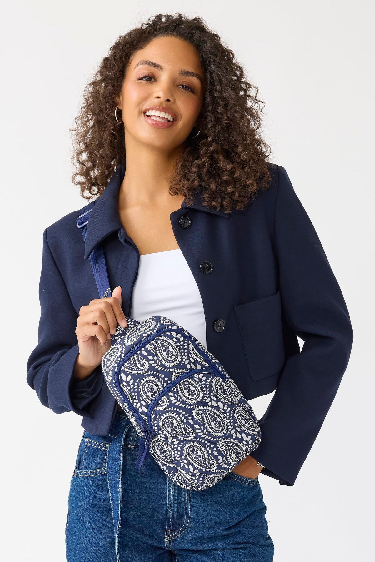 A woman with curly brown hair models a navy blue cropped jacket, blue jeans, and a white tank top, while carrying a small patterned crossbody bag with a blue strap.